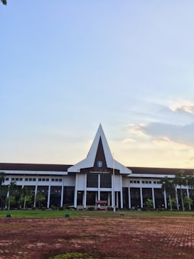 A large building with a pointed triangular roof stands in the center, surrounded by greenery and trees on either side. The building displays a crest above its main entrance, indicating an official or governmental function. The foreground features a paved area with patches of grass, and the sky above is clear with a few clouds, suggesting a peaceful setting.