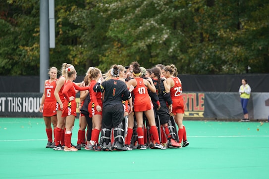 A group of female field hockey players in red uniforms gather in a huddle on a green field. They appear to be discussing or strategizing before a game or after a timeout. The team members are wearing protective gear, and some have numbers on their jerseys. The background shows a fence and trees, suggesting an outdoor sports complex.