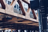 A metallic train travels on an elevated railway track supported by a riveted steel structure. The scene includes urban architecture in the background, suggesting a city environment. A traffic light and signage are also visible in the foreground, contributing to the urban setting.