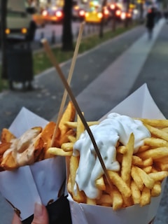 Two paper cones filled with fries, one topped with creamy white sauce and the other with a different sauce, held up by wooden sticks. They are held by someone on a blurred city street backdrop with lights and people in the distance.