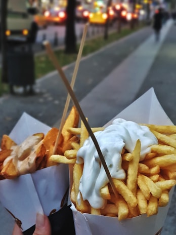 Two paper cones filled with fries, one topped with creamy white sauce and the other with a different sauce, held up by wooden sticks. They are held by someone on a blurred city street backdrop with lights and people in the distance.