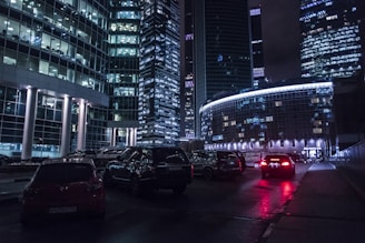 A nighttime cityscape featuring modern high-rise buildings with glass facades illuminated by artificial lights. The reflections on the windows create a mosaic of lights. Numerous cars are visible on the street, with headlights and taillights creating streaks of red and white.