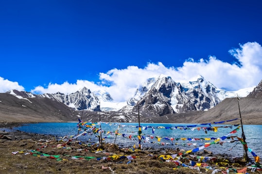 A serene mountain landscape featuring a calm blue lake in the foreground, surrounded by snow-capped peaks under a clear blue sky. Colorful prayer flags are draped prominently across the scene, adding vibrant contrast to the natural setting.