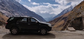 A black SUV is parked on a rough road amidst a mountainous landscape. Majestic mountains rise on either side, with some peaks covered in snow. The sky is partially cloudy, adding depth to the scene. The terrain appears rugged and remote, suggesting a sense of adventure.