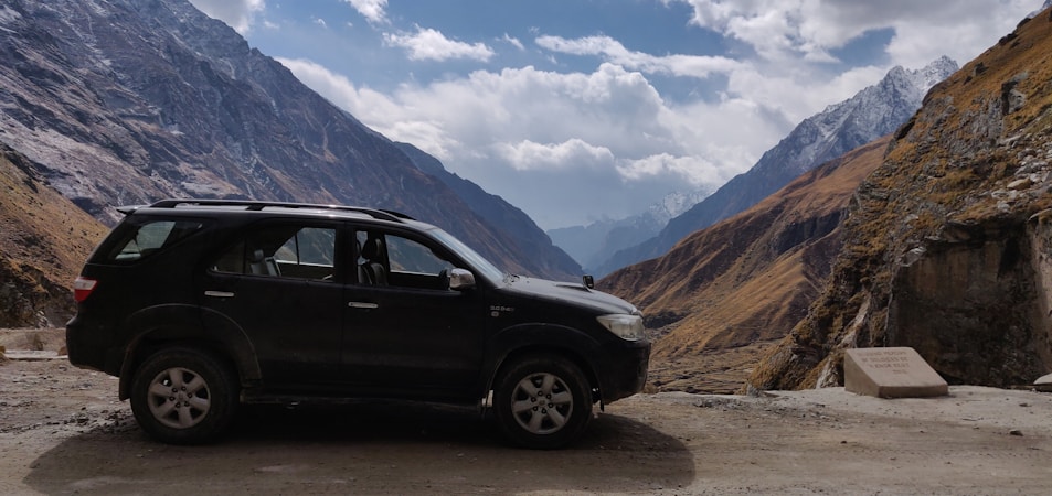 A black SUV is parked on a rough road amidst a mountainous landscape. Majestic mountains rise on either side, with some peaks covered in snow. The sky is partially cloudy, adding depth to the scene. The terrain appears rugged and remote, suggesting a sense of adventure.