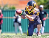A football player wearing a helmet and a sports uniform is on one knee, intently focusing on catching a football. The player is adorned with arm tattoos and wears a jersey with the number 33. In the background, other individuals can be seen standing near a green fence, out of focus.