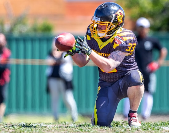 A football player wearing a helmet and a sports uniform is on one knee, intently focusing on catching a football. The player is adorned with arm tattoos and wears a jersey with the number 33. In the background, other individuals can be seen standing near a green fence, out of focus.