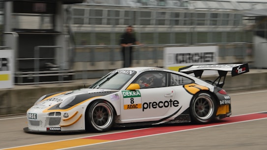 A white and black sports car is speeding on a race track. The car features sponsor logos, a large rear wing, and a sleek aerodynamic design. The environment includes barriers and blurred motion, indicating high speed. A person is visible near the pit area.