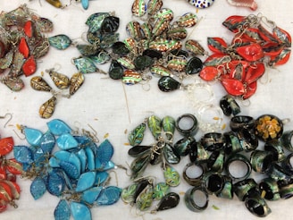 A collection of colorful handmade earrings and rings spread out on a white fabric background. The earrings are in various shapes and colors, including vibrant reds, blues, greens, and gold. Some earrings feature intricate patterns and details. A number of thick, dark green rings are also present.