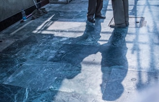 The image features two individuals with only their lower bodies visible. They are standing on a shiny, polished stone floor with visible reflections and shadows. There are cleaning supplies, including a mop and a bottle, placed against the wall on the left side of the image.