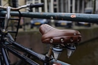 A close-up view of a bicycle seat with a brown leather cover attached to a black metal frame. The background features a blurred view of parked cars and buildings.