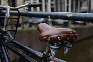 A close-up view of a bicycle seat with a brown leather cover attached to a black metal frame. The background features a blurred view of parked cars and buildings.