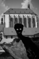 A silhouette of a human-like figure with hollow eyes and an open mouth stands prominently in the foreground, adding a mysterious and eerie vibe. In the background, there is a blurred image of a large, Gothic architectural structure, possibly a church, with pointed arches and a steep roof under a cloudy sky.