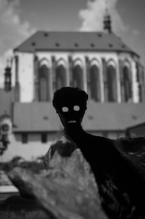 A silhouette of a human-like figure with hollow eyes and an open mouth stands prominently in the foreground, adding a mysterious and eerie vibe. In the background, there is a blurred image of a large, Gothic architectural structure, possibly a church, with pointed arches and a steep roof under a cloudy sky.