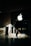 A person in business attire stands in front of a large glass window displaying a prominent white apple logo. Sunlight creates complex shadow patterns across the stone floor and walls, enhancing the contrast between light and dark areas.