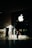 A person in business attire stands in front of a large glass window displaying a prominent white apple logo. Sunlight creates complex shadow patterns across the stone floor and walls, enhancing the contrast between light and dark areas.
