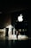 A person in business attire stands in front of a large glass window displaying a prominent white apple logo. Sunlight creates complex shadow patterns across the stone floor and walls, enhancing the contrast between light and dark areas.