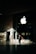 A person in business attire stands in front of a large glass window displaying a prominent white apple logo. Sunlight creates complex shadow patterns across the stone floor and walls, enhancing the contrast between light and dark areas.