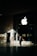 A person in business attire stands in front of a large glass window displaying a prominent white apple logo. Sunlight creates complex shadow patterns across the stone floor and walls, enhancing the contrast between light and dark areas.