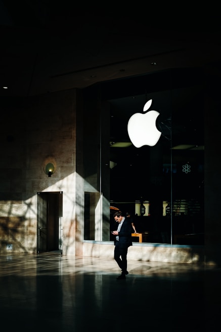 A person in business attire stands in front of a large glass window displaying a prominent white apple logo. Sunlight creates complex shadow patterns across the stone floor and walls, enhancing the contrast between light and dark areas.