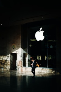A person in business attire stands in front of a large glass window displaying a prominent white apple logo. Sunlight creates complex shadow patterns across the stone floor and walls, enhancing the contrast between light and dark areas.