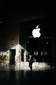 A person in business attire stands in front of a large glass window displaying a prominent white apple logo. Sunlight creates complex shadow patterns across the stone floor and walls, enhancing the contrast between light and dark areas.