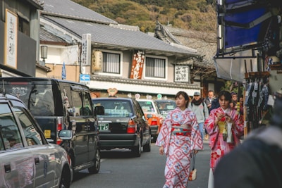 Two women dressed in colorful kimonos walk along a bustling street lined with parked cars and traditional Japanese buildings. Various signs in Japanese adorn the storefronts, and people can be seen casually strolling in the background.