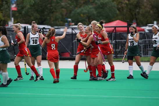 A group of female field hockey players wearing red and green uniforms are gathered on a green turf field. Some players appear to be celebrating, with smiles and gestures of camaraderie. Others are standing nearby with neutral expressions, holding field hockey sticks.