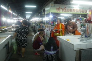 A market scene with fluorescent lighting and several individuals. A Buddhist monk in an orange robe holds a metal alms bowl. A man beside him appears to be assisting or accompanying him. People are kneeling or standing in front of the monk, likely engaged in a religious offering or prayer. Various stalls with goods, likely food items, line the sides of the walkway.