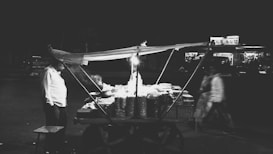 A street vendor cart is displayed at night, illuminated by a single light. The cart is filled with various goods and is covered by a makeshift canopy. A person stands near the cart, while another person walks by in the background. The scene is set against a dark street with dimly lit buildings in the background.
