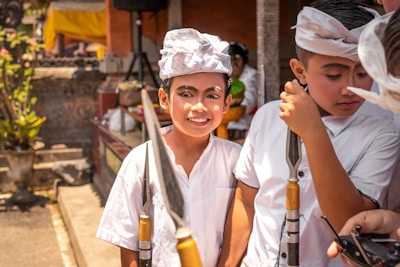 Two children dressed in white traditional attire with headscarves are smiling and interacting. They are holding ceremonial spears in an outdoor setting that appears to be a cultural event or festival.
