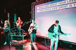 A band performs on stage with musicians playing guitars and a bass. The stage is equipped with microphones and amplifiers. The background features a large screen displaying lyrics of a song, illuminated by colorful stage lights.