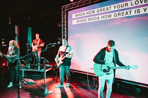 A band performs on stage with musicians playing guitars and a bass. The stage is equipped with microphones and amplifiers. The background features a large screen displaying lyrics of a song, illuminated by colorful stage lights.