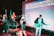 A band performs on stage with musicians playing guitars and a bass. The stage is equipped with microphones and amplifiers. The background features a large screen displaying lyrics of a song, illuminated by colorful stage lights.