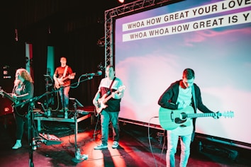 A band performs on stage with musicians playing guitars and a bass. The stage is equipped with microphones and amplifiers. The background features a large screen displaying lyrics of a song, illuminated by colorful stage lights.