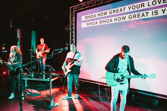 A band performs on stage with musicians playing guitars and a bass. The stage is equipped with microphones and amplifiers. The background features a large screen displaying lyrics of a song, illuminated by colorful stage lights.