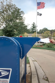 A hand is inserting an envelope into a blue United States Postal Service mailbox situated on a suburban street. In the background, an American flag and a POW/MIA flag are hoisted on a flagpole. There are trees and a stop sign visible, suggesting a calm neighborhood setting.