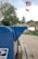 A hand is inserting an envelope into a blue United States Postal Service mailbox situated on a suburban street. In the background, an American flag and a POW/MIA flag are hoisted on a flagpole. There are trees and a stop sign visible, suggesting a calm neighborhood setting.