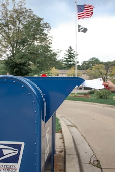 A hand is inserting an envelope into a blue United States Postal Service mailbox situated on a suburban street. In the background, an American flag and a POW/MIA flag are hoisted on a flagpole. There are trees and a stop sign visible, suggesting a calm neighborhood setting.