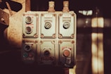 An industrial control panel featuring several switches and dials, each labeled with instructions like 'can stop' and 'job run'. The panel is metallic and weathered, suggesting it is used in a rugged environment. Soft sunlight casts shadows across the panel giving it a vintage appearance.