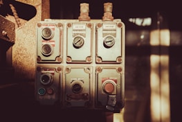 An industrial control panel featuring several switches and dials, each labeled with instructions like 'can stop' and 'job run'. The panel is metallic and weathered, suggesting it is used in a rugged environment. Soft sunlight casts shadows across the panel giving it a vintage appearance.