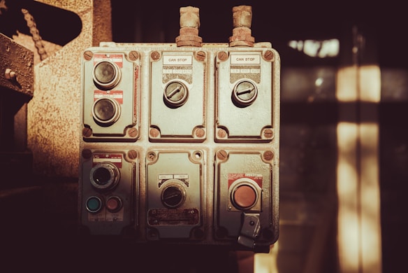 An industrial control panel featuring several switches and dials, each labeled with instructions like 'can stop' and 'job run'. The panel is metallic and weathered, suggesting it is used in a rugged environment. Soft sunlight casts shadows across the panel giving it a vintage appearance.