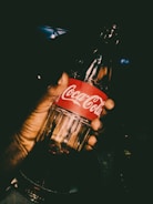 A hand is holding a glass bottle of Coca-Cola with the logo clearly visible on a red label. The lighting is dramatic, creating a high contrast and focusing attention on the bottle.