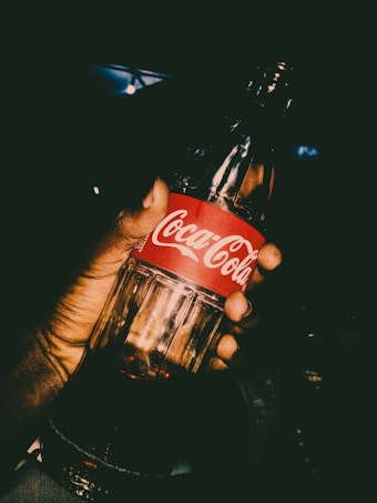 A hand is holding a glass bottle of Coca-Cola with the logo clearly visible on a red label. The lighting is dramatic, creating a high contrast and focusing attention on the bottle.