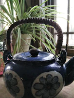A bright ceramic teapot steaming gently beside a window with sunlight filtering through green plants.