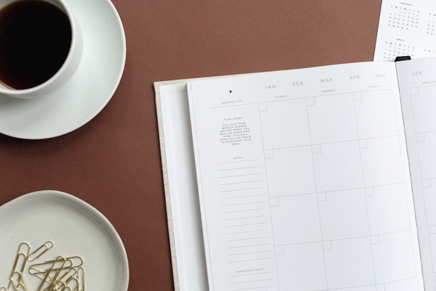 An open planner rests on a brown surface. It displays a calendar layout with sections for each day of the week across multiple months. On the left side, a cup containing a dark beverage is placed on a saucer. Nearby, there is a small dish filled with gold paperclips. A partial view of another calendar page can be seen in the background.