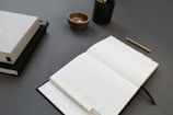 An open planner rests on a dark surface alongside a closed book, a small wooden bowl, a black container with pencils, and a gold pen laid neatly.