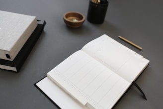 An open planner rests on a dark surface alongside a closed book, a small wooden bowl, a black container with pencils, and a gold pen laid neatly.