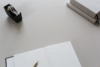 A clean and minimalist workspace featuring an open planner with a gold pen resting on it, a black tape dispenser with metallic components, and two closed gray books stacked on one another.