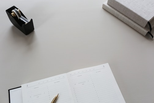 A clean and minimalist workspace featuring an open planner with a gold pen resting on it, a black tape dispenser with metallic components, and two closed gray books stacked on one another.